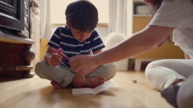 Genç bir çocuk kırmızı bir kalemle çizim aktivitesine konsantre olurken, annesi de evlerinin zemininde odaklanmış bir ev okulu anını vurguluyor.