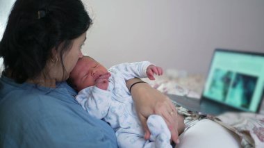 Anne çoklu görevi, kucağında yeni doğmuş bir bebeği dizüstü bilgisayarla rahat yatak odası ortamında çalışırken tutmak, modern ebeveynlik, iş hayatı dengesi ve anne bakımı örneği vermek, çevrimiçi aktivitelerde birlikte olmak.