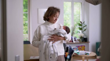 Büyük anne parlak bir evde duruyor, yeni doğan torununu yumuşak bir battaniyeye sarıp kucaklıyor, sıcaklığı, ilgiyi ve nesiller arası bağı ön plana çıkaran sevgi dolu bir an.