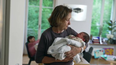 Yaşlı bir kadın, yumuşak bir battaniyeye sarılı yeni doğan torununa sevgiyle bakıyor. Çağdaş bir evde. Aile sevgisini, ilgiyi ve nesiller arasındaki bağı sembolize ediyor.