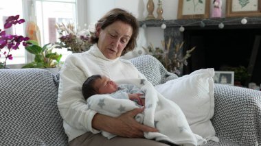 Büyükanne kadın yeni doğmuş bebeği rahat oturma odasında kucaklıyor, sıcak ve sevgi dolu bir aile anı, şefkatli kucaklama, nesiller arası bağ, ev içi huzur verici sahne