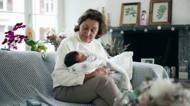 Yaşlı bir kadın yeni doğmuş bebeği rahat bir oturma odasında kucaklıyor, sıcak ve sevgi dolu bir aile anı, büyükannenin şefkatli kucaklaması, nesiller arası bağ kurması, ev içleri.