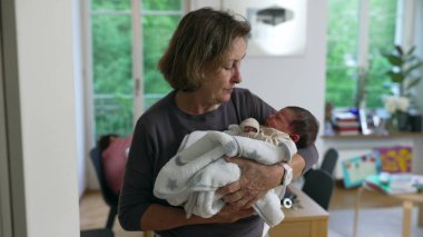 Yaşlı bir kadın, yumuşak bir battaniyeye sarılı yeni doğan torununa sevgiyle bakıyor. Çağdaş bir evde. Aile sevgisini, ilgiyi ve nesiller arasındaki bağı sembolize ediyor.