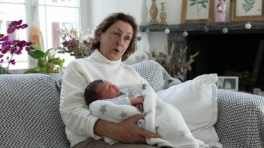 Büyükanne kadın yeni doğmuş bebeği rahat oturma odasında kucaklıyor, sıcak ve sevgi dolu bir aile anı, şefkatli kucaklama, nesiller arası bağ, ev içi huzur verici sahne