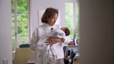 Büyük anne parlak bir evde duruyor, yeni doğan torununu yumuşak bir battaniyeye sarıp kucaklıyor, sıcaklığı, ilgiyi ve nesiller arası bağı ön plana çıkaran sevgi dolu bir an.