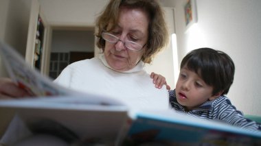 Büyükannesi, torununa renkli bir kitap okuyor. Torunu da onu dinlemek için eğiliyor. Büyükanne gözlük ve beyaz bir kazak giyerken, çizgili gömlekli çocuk kitaba dikkatle bakıyor.