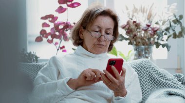 Evde akıllı telefon kullanan yaşlı bir kadın, gözlük takıyor ve ekrana odaklanıyor, yaşlıların modern yaşam tarzını ve dijital bağlantılarını yakalıyor, bağlantı ve teknoloji kullanımını vurguluyor
