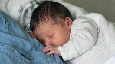 Yeni doğmuş bir bebeğin, yumuşak beyaz bir battaniyeye sarılı bir ebeveynin göğsünde huzurlu bir şekilde uyuması. Bebeğin dinlenme sıcaklığı ve güvenliği.