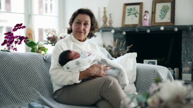 Yaşlı bir kadın yeni doğmuş bebeği rahat bir oturma odasında kucaklıyor, sıcak ve sevgi dolu bir aile anı, büyükannenin şefkatli kucaklaması, nesiller arası bağ kurması, ev içleri.