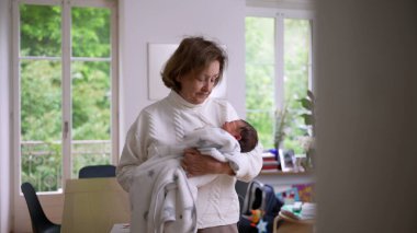 Büyük anne parlak bir evde duruyor, yeni doğan torununu yumuşak bir battaniyeye sarıp kucaklıyor, sıcaklığı, ilgiyi ve nesiller arası bağı ön plana çıkaran sevgi dolu bir an.