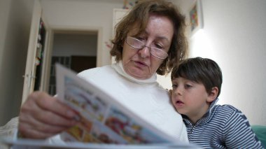 Büyükannesi, torununa renkli bir kitap okuyor. Torunu da onu dinlemek için eğiliyor. Büyükanne gözlük ve beyaz bir kazak giyerken, çizgili gömlekli çocuk kitaba dikkatle bakıyor.