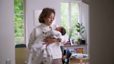 Büyük anne parlak bir evde duruyor, yeni doğan torununu yumuşak bir battaniyeye sarıp kucaklıyor, sıcaklığı, ilgiyi ve nesiller arası bağı ön plana çıkaran sevgi dolu bir an.