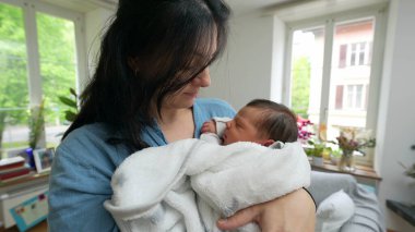 Anne beşikte, beyaz battaniyeye sarılı, modern oturma odasında, sevgiyle çocuğa bakıyor.