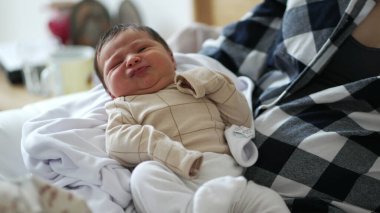 Yeni doğan bebek annesinin kucağında hafif bir gülümsemeyle, çocukluğun neşeli ve sevimli bir anını yakalıyor, anne ve çocuğu arasındaki güçlü bağı ve sevgiyi vurguluyor.