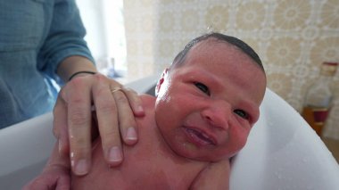 Banyo sırasında yeni doğan bebek, ebeveyn tarafından destekleniyor, gözler açık, cilt üzerinde su, erken bakım ve bağlanma anları, nezaket ve sevgiyi vurgulayarak,