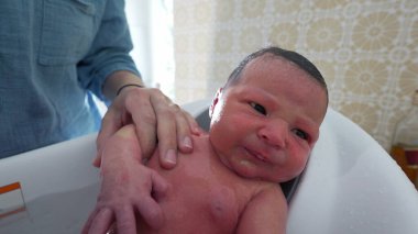 Banyo sırasında yeni doğan bebek, ebeveyn tarafından destekleniyor, gözler açık, cilt üzerinde su, erken bakım ve bağlanma anları, nezaket ve sevgiyi vurgulayarak,