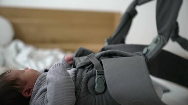 Anne, bebek taşıyıcı kayışları ayarlar, gri örgü süveter içinde yeni doğan bebeği korur, samimi bir bağ kurma anı, güvenliği ve rahatlığı sağlar, ebeveynliği sağlar, bebeğe bakar, dışarı çıkmaya hazırlanır.