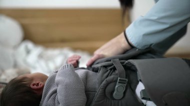 Anne, bebek taşıyıcı kayışları ayarlar, gri örgü süveter içinde yeni doğan bebeği korur, samimi bir bağ kurma anı, güvenliği ve rahatlığı sağlar, ebeveynliği sağlar, bebeğe bakar, dışarı çıkmaya hazırlanır.