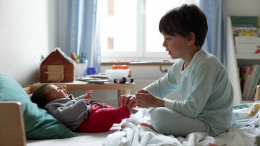 Büyük kardeş ve bebek, samimi bir yatakta, oyuncaklarla çevrili, yakın bağlarını ve sevgi dolu ilişkilerini vurgulayan güneşli bir odada, hassas bir anın tadını çıkarırlar.