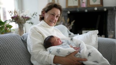 Büyükanne yeni doğmuş bebeği battaniyeye sardı, hassas ve samimi aile anı, ev ortamında bakım, çok nesilli bağ, rahat oturma odası, sevgi dolu sahne