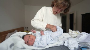 Büyükannem yeni doğan bebeklere beyaz tulum giydirir. Bebek yumuşak bir battaniyeye uzanır, ağzı şaşkınlıkla açılır, erken yaşlarda kuşak bağının ve şefkatin yürek ısıtan anını yakalar.