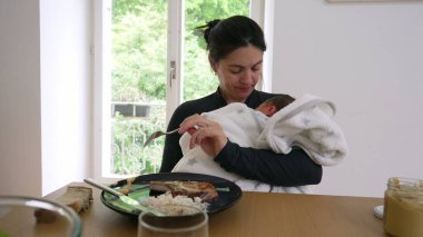 Anne yemek yerken yeni doğmuş bebeği kucağında tutuyor. Bakım ve kişisel ihtiyaçları dengelemek, erken anneliğin çoklu görev doğasını vurgulamak