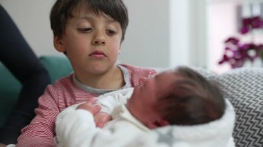 Küçük çocuk, yeni doğan kardeşini nazikçe kucağında tutuyor, annesi yakınlarda, aile dinamiklerini keşfediyor, besleniyor, kardeş bağı kuruyor, ev içinde samimi bir ortam, duygusal bir bağ kuruyor.