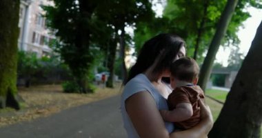 Anne, bebeğini ağaçlarla kaplı bir caddede kucaklıyor, derin sevgi ve özen gösteriyor, arka planda kentsel çevre ve yapraklı ağaçlar, huzurlu bir aile bağı anı.