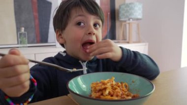 Spagettiyi çatalla çeviren bir çocuk, yemeğe yoğun bir şekilde odaklanırken onu sıkıca kavrıyor, tanıdık bir ev ortamında kendini besleme çabasını ve bağlılığını vurguluyor