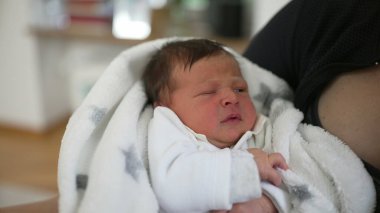 Yeni doğmuş bebek beyaz bir battaniyeye sarılmış, anne ve babası tarafından tutulan, hayatın ilk anlarını gözler önüne seren, aile sevgisi, bakım ve korunma, rahat bir ev ortamında, masumiyet.