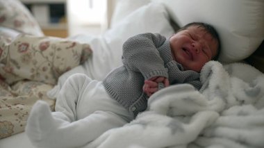 Yeni doğmuş bebek yatakta ağlıyor, gri örgü süveter giyiyor, beyaz pantolon giyiyor, yumuşak battaniyeye sarılmış, duygularını ifade ediyor, hayatın erken evreleri, bebek ağlamasının hassas anı.