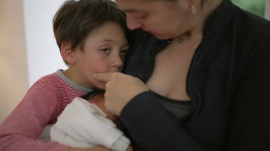 Bir anne kucağında yeni doğmuş bir bebeği kollarında tutarken küçük oğlunu teselli eder. Şefkat ve sevgi dolu bir anı paylaşır. Günlük kıyafetler giyer, duygusal bağ kurar, ebeveynlik yapar, aile desteğini paylaşır.