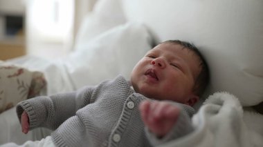 Yeni doğmuş bebek yatağa uzanıyor, gri örgü süveter giyiyor, beyaz pantolon giyiyor, rahat bir battaniyeye sarılmış, küçük eller ve ayaklar görünür, bebek dinleniyor, hayatın ilk anlarını yakalıyor, huzurlu bir sahne.