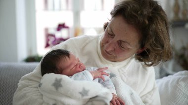 Yaşlı bir kadın sevgi dolu bir şekilde yeni doğan bebeğe bakıyor, içten bir aile anı, nesiller arası bağ, büyükanne ve torun, rahat bir oturma odası, ev ortamını besleme