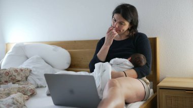 Anne işi dengelemek ve ebeveynlik, yeni doğmuş bebeği yatakta dizüstü bilgisayar kullanırken tutmak, modern aile dinamiklerini, çoklu görevleri ve evde çalışan ebeveynlerin günlük zorluklarını temsil etmek.
