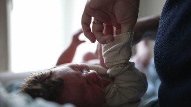 Anne yeni doğmuş bebeği giydiriyor, nazik eller tişörtü kaldırıyor, sevgi ve ilgi gösteriyor, yakınlık kurma anı, ebeveynliğin ilk aşamaları, hassas dokunuşlar ve yetiştirme sahnesi.