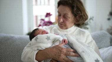 Yaşlı bir kadın sevgi dolu bir şekilde yeni doğan bebeğe bakıyor, içten bir aile anı, nesiller arası bağ, büyükanne ve torun, rahat bir oturma odası, ev ortamını besleme