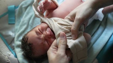 Anne yeni doğmuş bebeği giydiriyor, tişörtünü kafasının üstüne kaldırıyor, hassas bir ilgi anı, ebeveyn sevgisinin ve ilgisinin vurgulanması, bebek bakımının erken aşamaları, samimi ve besleyici sahneler.