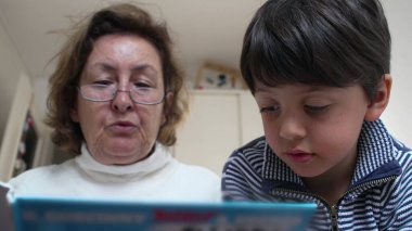 Gözlüklü büyükanne özenli torununa kitap okuyor. Aile bağlarının ve ortak öğrenmenin önemini vurgulayan odaklanmış ifadeler ve ilgi çekici hikaye anlatma oturumu