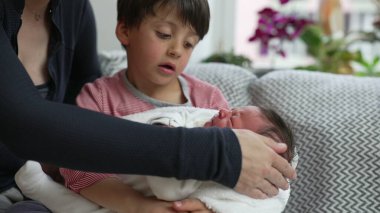 Anne ve küçük çocuk birlikte oturuyor, yeni doğan kardeşlerini beyaz battaniyeye sarıyorlar, aile bağlarını, kardeş sevgisini, aile sevgisini, ev ortamını, sevgi dolu aile anını yaşıyorlar.