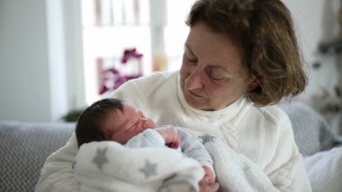Yaşlı bir kadın sevgi dolu bir şekilde yeni doğan bebeğe bakıyor, içten bir aile anı, nesiller arası bağ, büyükanne ve torun, rahat bir oturma odası, ev ortamını besleme