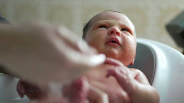 Banyo sırasında yeni doğan bebek, düşünceli bir ifadeyle etrafına bakıyor, merak ve farkındalık anı, anne tarafından sağlanan bakımı yansıtıyor.