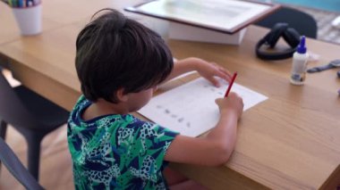 Bir çocuk ahşap bir masada kağıt üzerinde bir kalem kullanarak derinden çizime odaklanır. Sessiz bir an yaratıcılığı ve konsantrasyonu yansıtır. Çocuk sanatsal aktivitelerine dalmıştır.