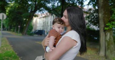 Anne, bebeğini ağaçlarla kaplı bir şehir yolunda tutuyor. Etrafı binalar ve yeşilliklerle çevrili çocuğuna sevgiyle bakıyor. Sakin ve besleyici bir atmosferi var.