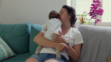 Anne sıcak bir kanepede otururken bebeğini nazikçe tutuyor. Şefkatli an, anne ve çocuğu arasındaki derin sevgi ve bağın altını çizer.