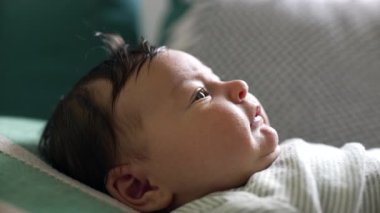 Yumuşak yastığa uzanmış yeni doğmuş bir bebeğin yan profili, gözleri tamamen açık ve çevreyi gözlemliyor, sakin, kapalı bir ortamda bir merak ve masumiyet anı.