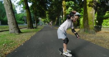 Çocuk kaykay yapıyor ağaçlarla kaplı bir yolda, miğfer ve koruyucu ekipmanlarla donatılmış, tahtada dengelenmiş, açık havada güneşli bir günün tadını çıkarıyor, yeşillik ve doğa ile çevrili.