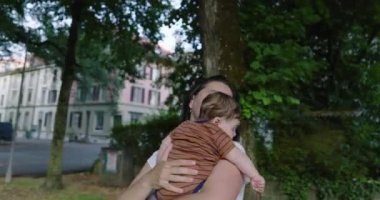 Anne, ağaçlarla çevrili bir şehir yolunda bebeğini tutarken huzur ve sevgi dolu bir ortamda konut binaları ile birlikte bir yere bakıyor.