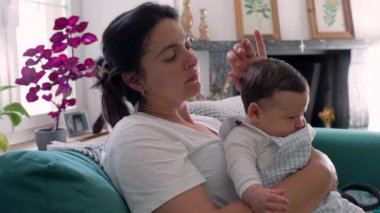 Anne nazikçe bebeğini kucağında tutuyor, her ikisi de bir an için aşağıya bakıyor, ev dekorasyonu ve mor bitkilerle çevrili, aşkı ve birlikteliği simgeliyor.