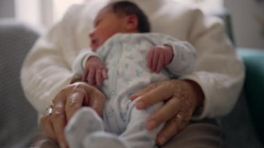 Yaşlı bir kadının elleri, yumuşak tulum giymiş yeni doğmuş bir bebeği kucaklıyor. Nazik bir ilgi, sevgi ve nesiller arasındaki hassas bağı yakalıyor.